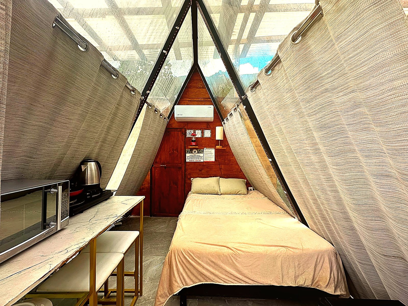 Interior de ATENEA con cama matrimonial, cocineta equipada, climatización y cortinas texturizadas bajo el ventanal A-frame